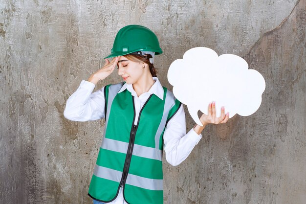 Free photo female engineer in green uniform and helmet holding a cloud shape info board and looks tired and sleepy.