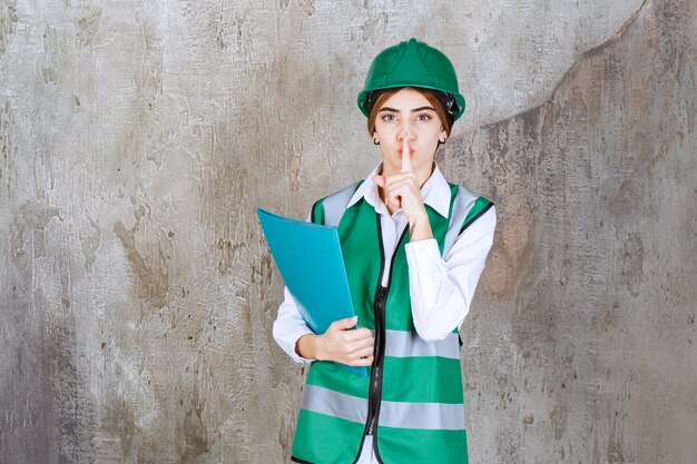 緑の制服とヘルメットの女性エンジニアは、緑のプロジェクトフォルダーを保持し、沈黙を求めています。