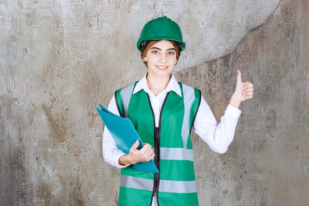 Female engineer in green uniform and helmet holding a green project folder and showing positive hand sign.