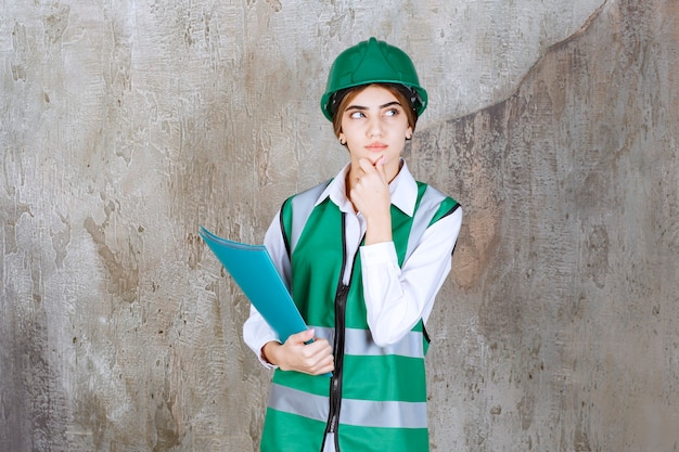 Женщина-инженер в зеленой форме и шлеме держит зеленую папку проекта, думает и анализирует