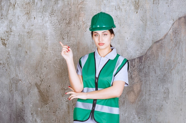 Женский инженер в зеленой форме и шлеме показывает левую сторону с эмоциями.