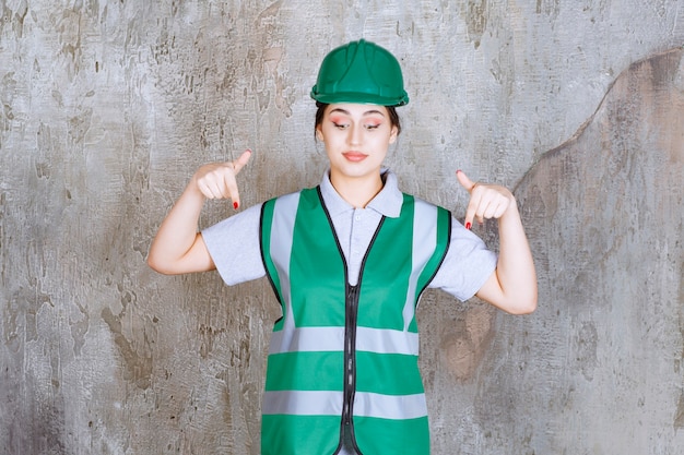 Foto gratuita ingegnere femminile in uniforme verde e casco che mostra qualcosa dietro.
