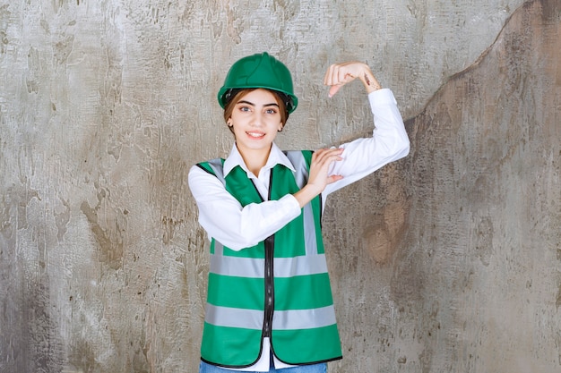 Foto gratuita ingegnere femminile in uniforme verde e casco in piedi sul muro di cemento e dimostrando i muscoli del braccio.