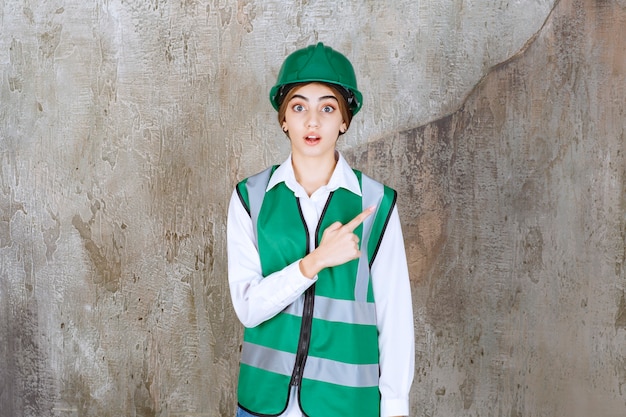 Female engineer in green uniform and helmet standing on concrete wall and showing right side.