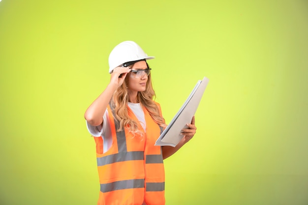 Female engineer in helmet and gear holding project plan and wearing her glasses.
