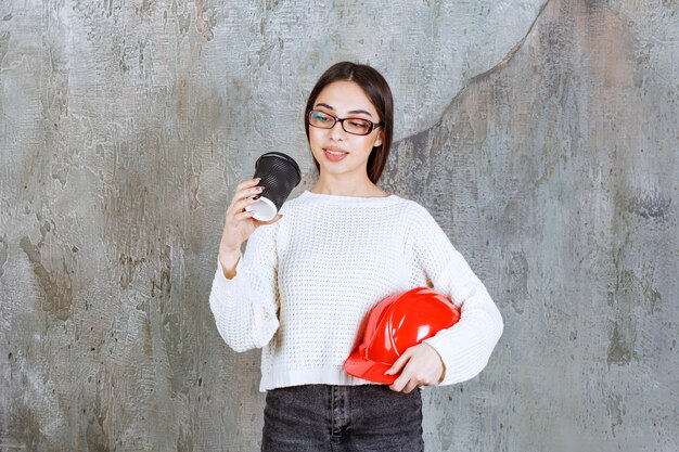 Female engineer holding a red helmet and a black disposable cup of drink and looks thoughtful.