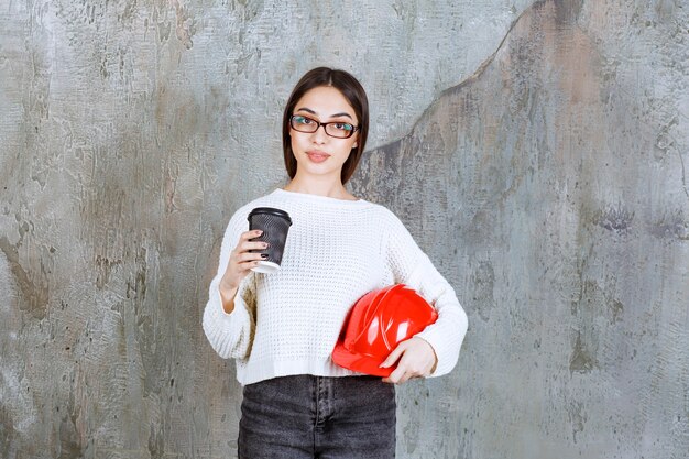 Female engineer holding a red helmet and a black disposable cup of drink.