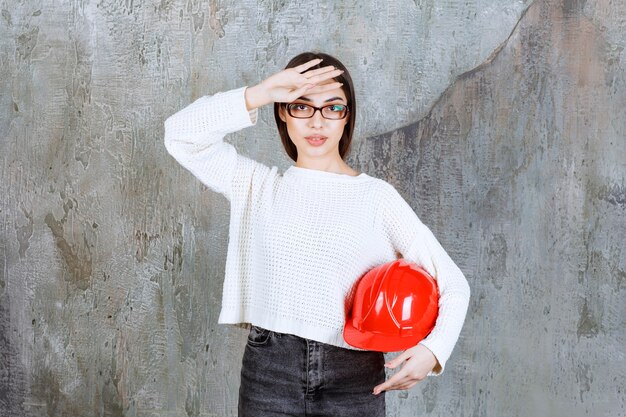 Женщина-инженер держит красный шлем и выглядит усталой или у нее болит голова.