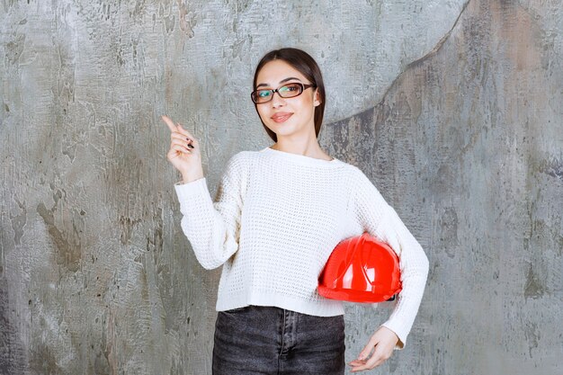 Female engineer holding a red helmet and showing something around.