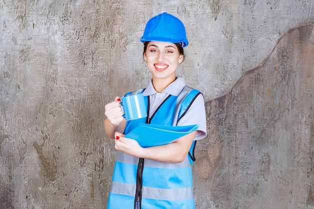 Женщина-инженер в синей форме и шлеме держит чашку синего чая и синюю папку с отчетом и предлагает напиток своему коллеге. Бесплатные Фотографии