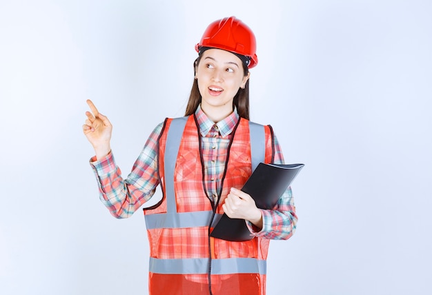 Female engineer in red helmet holding a black project plan and having a good idea.