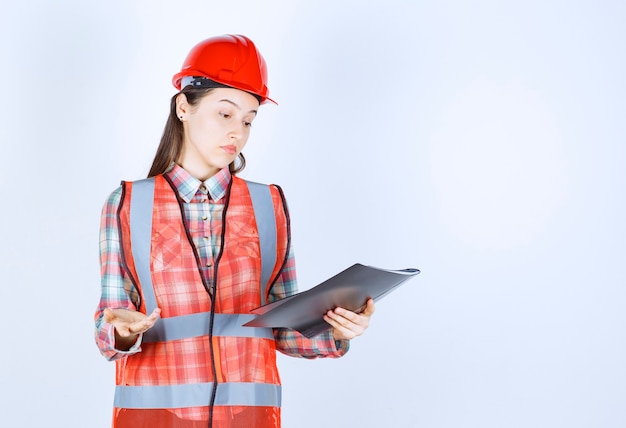 Free photo female engineer in red helmet holding a black project plan and looks confused.