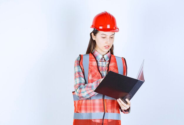 Female engineer in red helmet holding a black project plan.