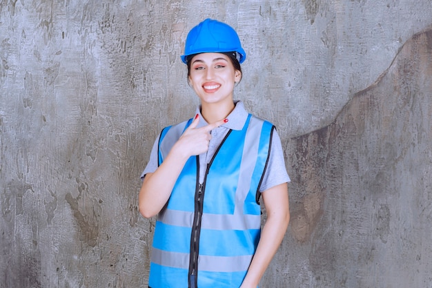 Foto gratuita ingegnere femminile che indossa casco e attrezzi blu e mostra qualcosa a destra.