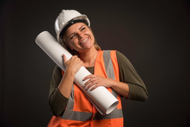 Женский инженер с белым шлемом держит план проекта и выглядит счастливым.