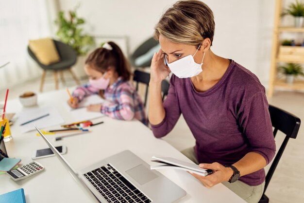 Female entrepreneur working on laptop while her daughter is doing homework during coronavirus lockdown