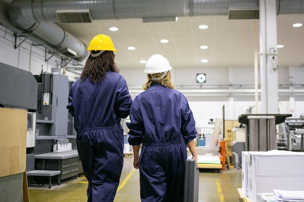 Free photo female factory workers in hardhats and overalls walking on plant floor and talking, carrying toolkit box