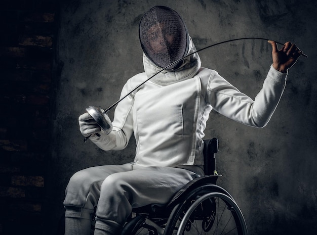 Female fencer in wheelchair with safety mask of a face, holding rapier on grey background.