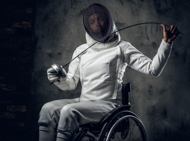 Женщина-фехтовальщица в инвалидной коляске с защитной маской на лице, держащая рапиру на сером фоне.