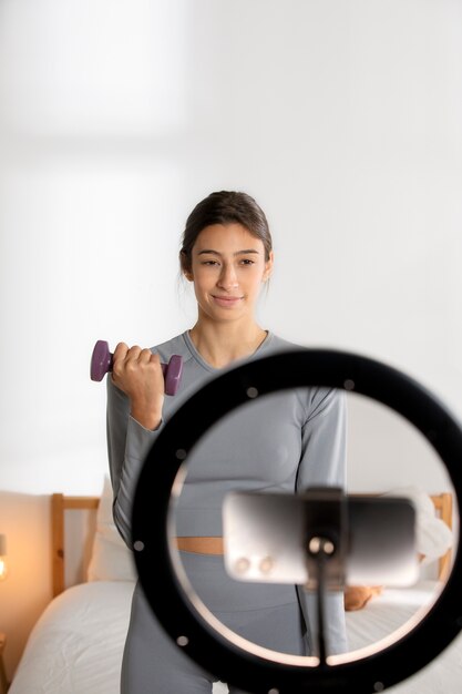 Female fitness instructor recording herself lifting weights at home with smartphone and ring light