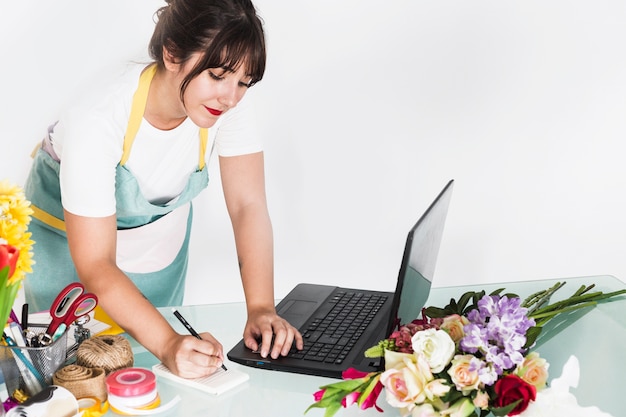 Female florist writing notes on notepad with laptop on desk