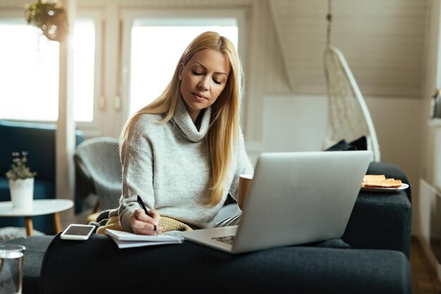 Female freelance worker writing in notebook while working on a computer at home