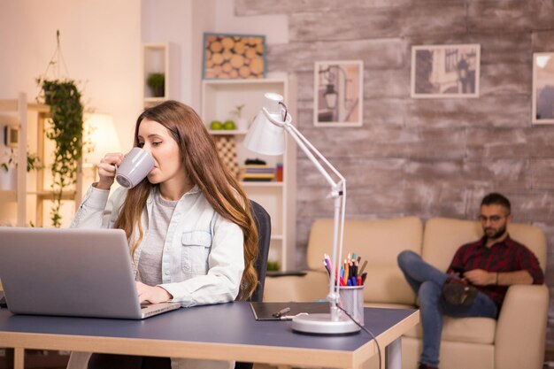 自宅で仕事をしながらラップトップで作業しながらコーヒーを楽しんでいる女性のフリーランサー。バックグラウンドでソファでリラックスした彼氏。