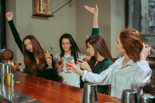 Free photo female friends having a drinks at bar. they are sitting at a wooden table with cocktails. they are clinking glasses