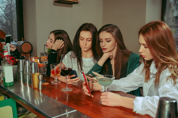 Female friends having a drinks at bar. They are sitting at a wooden table with cocktails. They are wearing casual clothes.