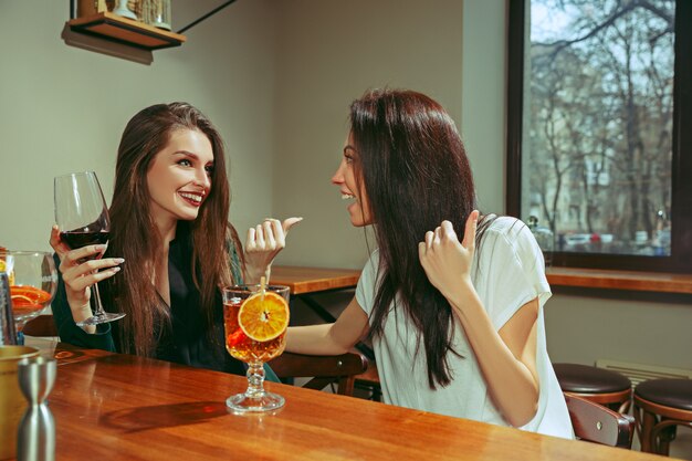 Female friends having a drinks at bar. They are sitting at a wooden table with cocktails.