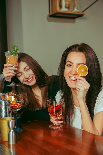 バーで飲み物を飲んでいる女性の友人。彼らはカクテルを片手に木製のテーブルに座っています。
