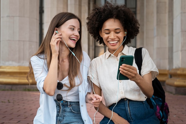 Female friends listening to music on earphones from smartphone outdoors