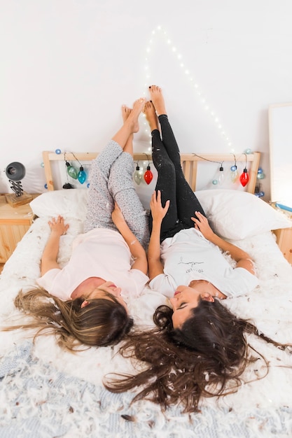 Female friends lying on messy bed with her legs up leaning on wall in bedroom