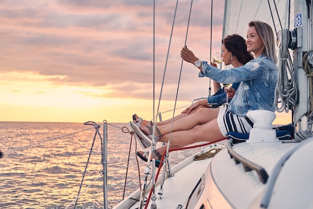 Female friends relaxing on the yacht with glasses of wine in the hands, during sunset on the high seas.