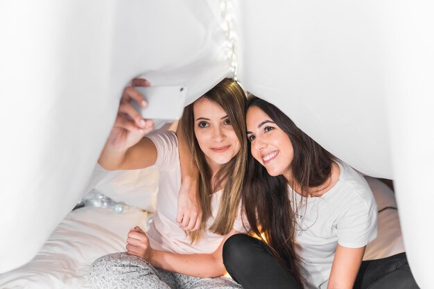 Female friends sitting on bed taking selfie on smartphone under the curtain