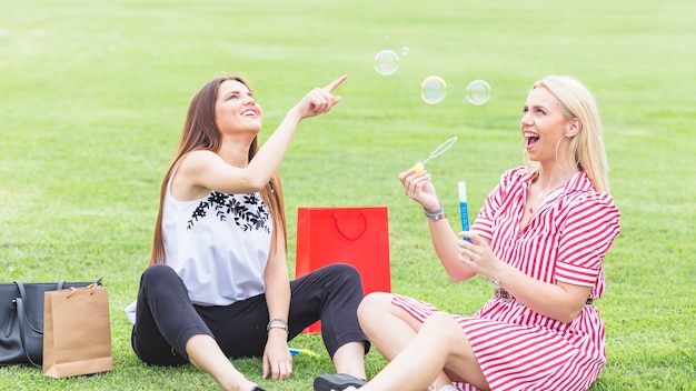 Female friends sitting on lawn playing with bubbles