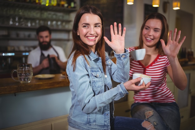 Female friends waving hands while having a cup of coffee
