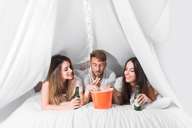 Amici femminili con uomo disteso sul letto, godendo di mangiare popcorn