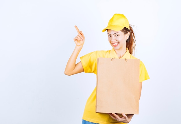Девушка в желтой форме держит картонную хозяйственную сумку и указывает куда-то.