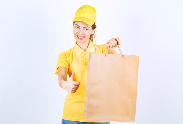 Female girl in yellow uniform holding a cardboard shopping bag and showing thumb up. 