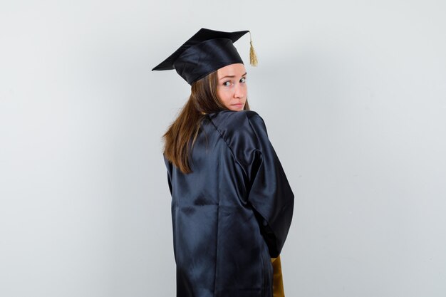 Female graduate in academic dress looking at camera over her shoulder and looking cute , back view.
