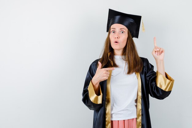 Female graduate in academic dress pointing up and right and looking puzzled , front view.