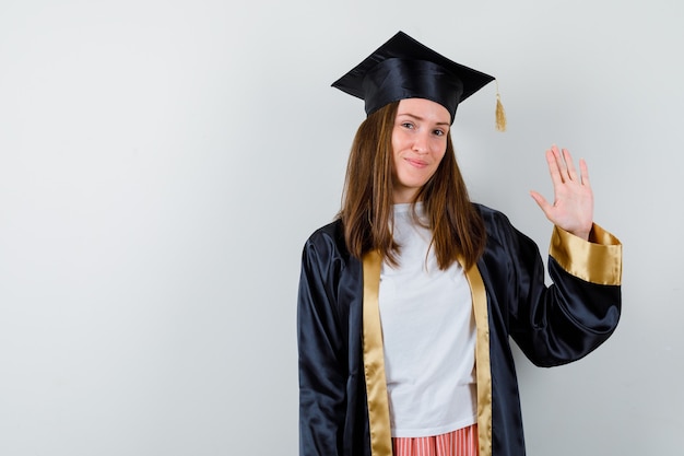 Free photo female graduate in academic dress waving hand to say goodbye and looking jolly , front view.