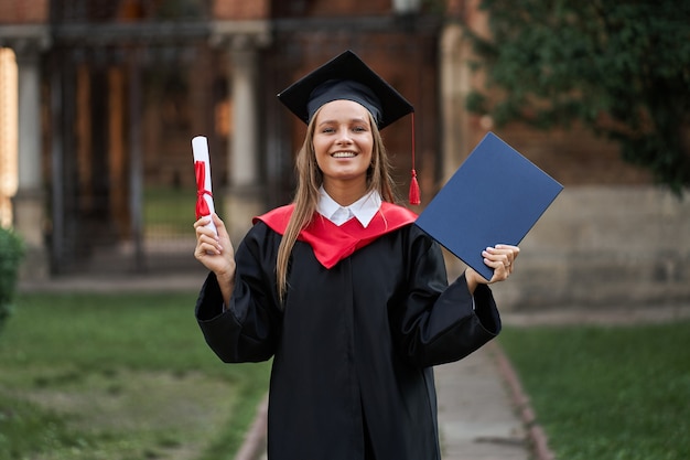 キャンパス内で卒業証書を手にした卒業ローブを着た女性卒業生。