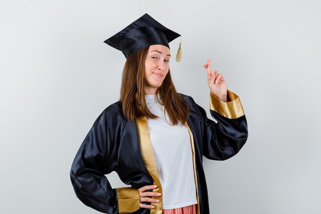 Female graduate keeping fingers crossed in academic dress and looking happy , front view.