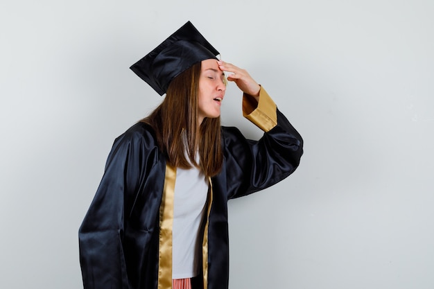 Free photo female graduate keeping hand on head in academic dress and looking annoyed , front view.