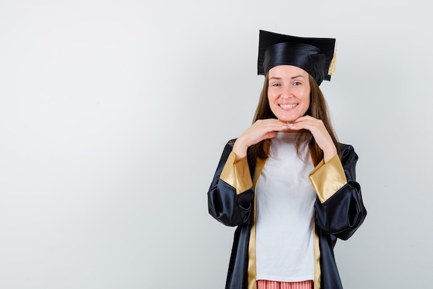 Foto gratuita laureato femminile che tiene le mani sotto il mento in abito accademico e sembra allegro. vista frontale.