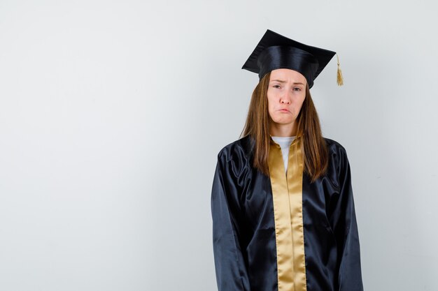 Female graduate looking at camera, curving lips, frowning face in academic dress and looking sad. front view.