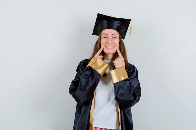 Free photo female graduate pointing at her teeth in academic dress and looking cheerful. front view.