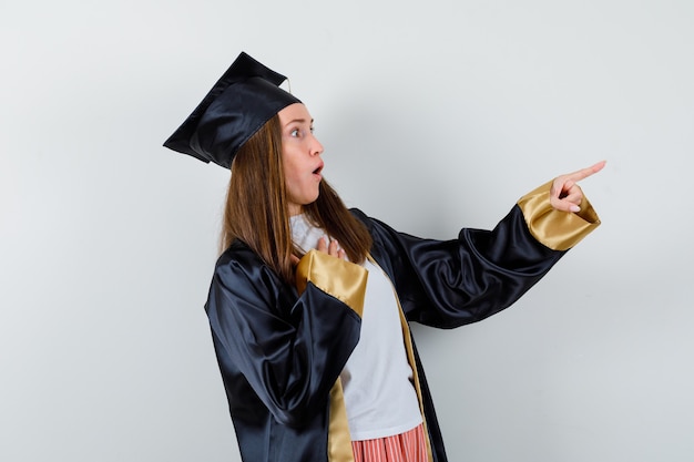 Foto gratuita laureato femminile che indica a destra in abito accademico e che sembra spaventato, vista frontale.